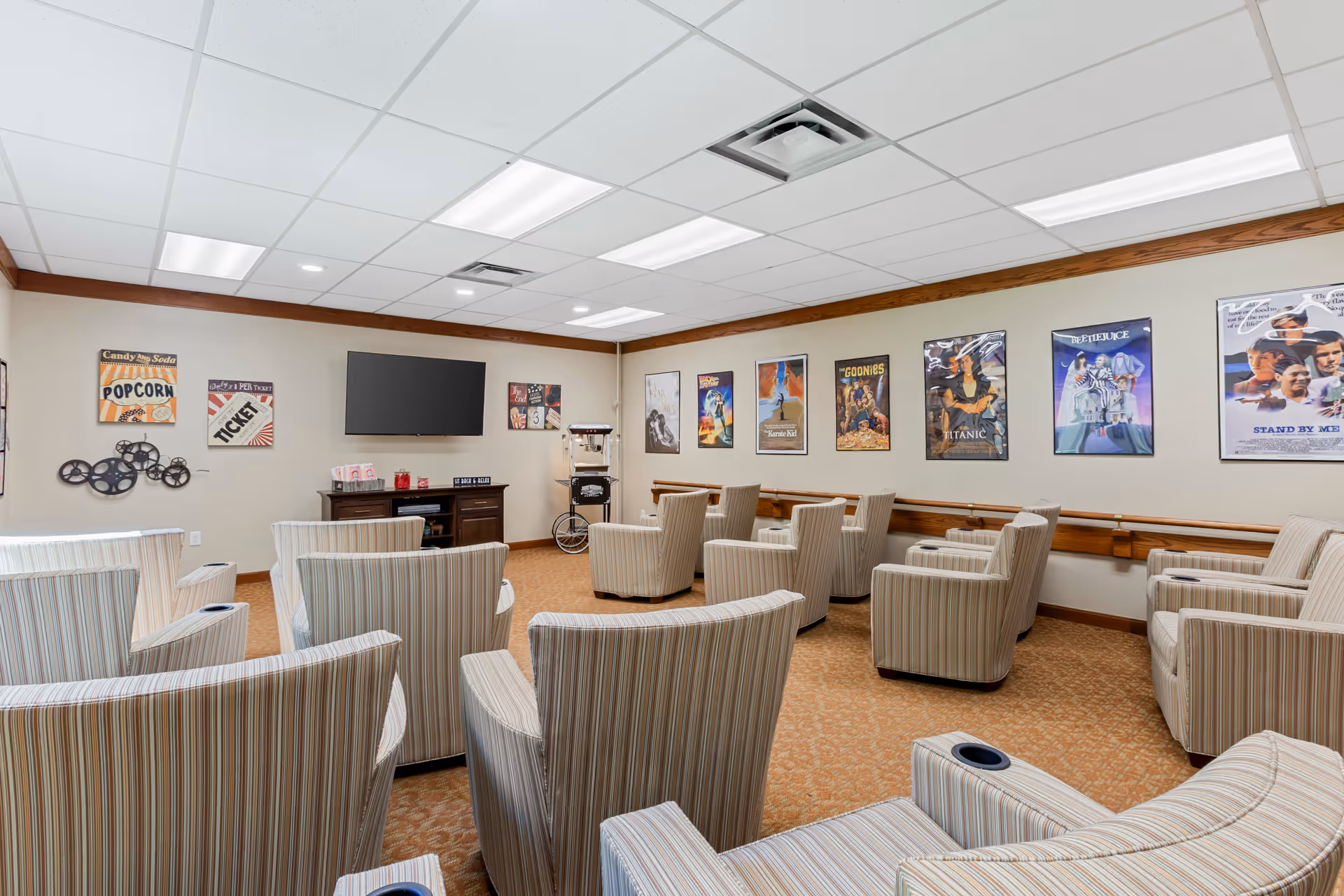 A cozy movie theater room with multiple striped armchairs arranged in rows facing a wall-mounted flat screen TV. The walls are decorated with vintage movie posters and popcorn-themed signs. There is a popcorn machine on a stand near the TV, and the room has a carpeted floor and a drop ceiling with fluorescent lights.