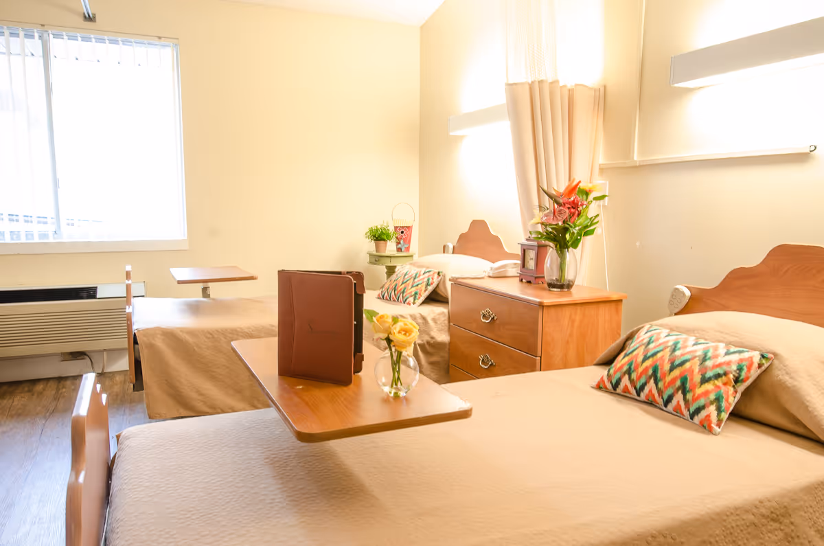 A bright room with two single beds, each with a colorful patterned pillow and beige bedspread. Between the beds is a wooden nightstand with a vase of flowers and a small clock. Each bed has an overbed table, one with a small vase of yellow flowers and a closed book or folder. A window with vertical blinds lets in natural light, and the walls are painted a soft yellow.
