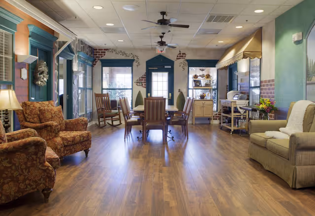 A cozy and well-lit common area with wooden flooring, featuring a round table with chairs in the center, two patterned armchairs on the left, and a cushioned sofa with a throw blanket on the right. The room has large windows with green trim, ceiling fans, and decorative elements including a wreath and a small cabinet with items on display.