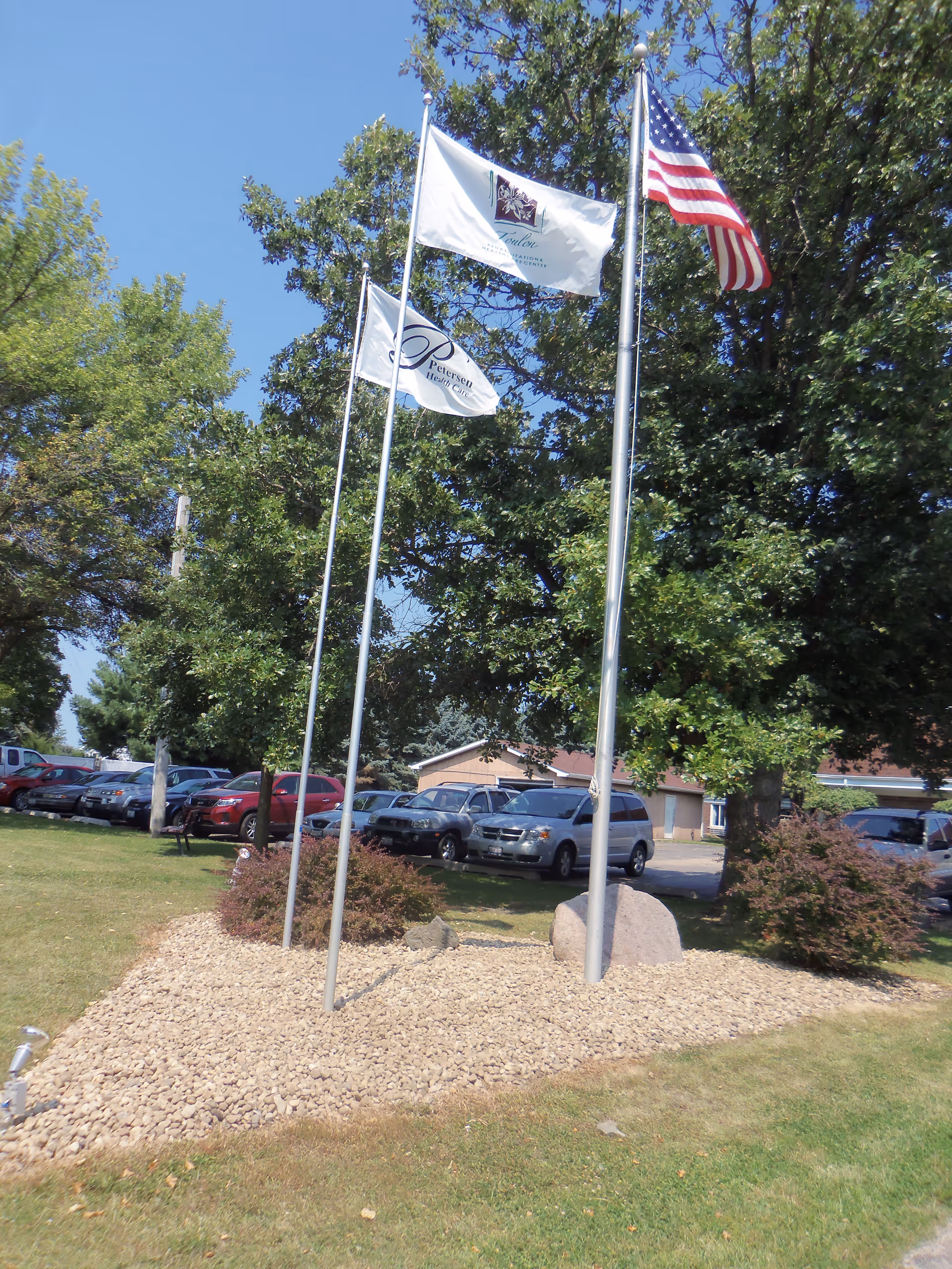 Three flagpoles with flags flying, including an American flag and two white flags with logos, set in a landscaped area with rocks and bushes. Behind the flags are trees and parked cars in a parking lot under a clear blue sky.