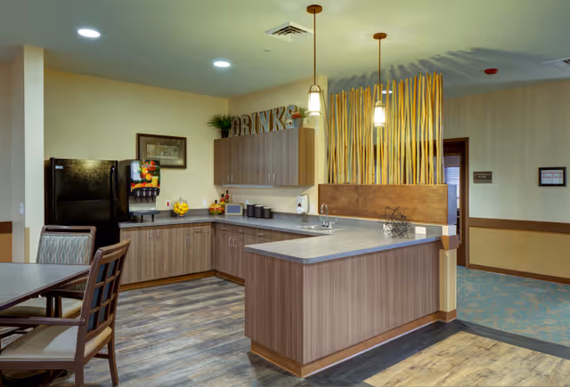 A modern kitchenette area with wooden cabinets, a black refrigerator, a drink dispenser, a microwave, and a counter with a sink. There are two pendant lights hanging above the counter and decorative bamboo sticks behind the counter. A dining table with chairs is partially visible on the left side.