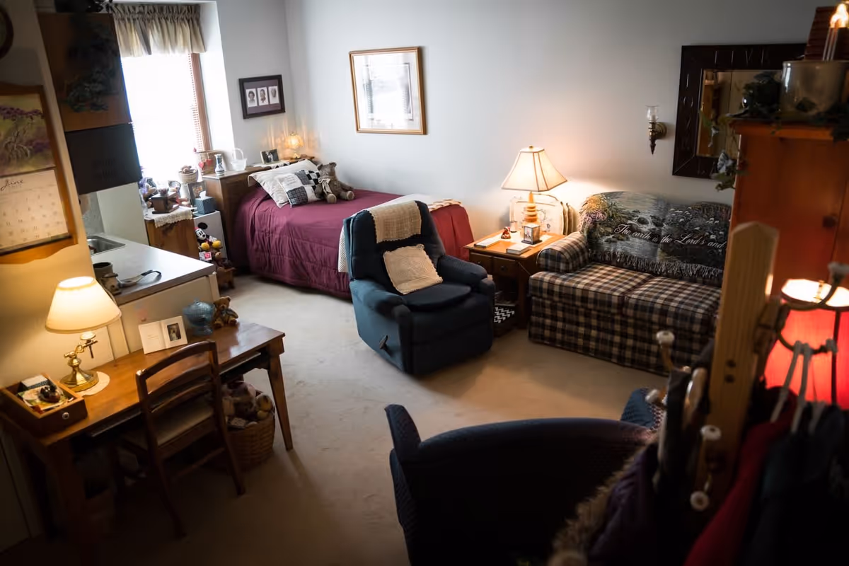 A cozy senior living room featuring a single bed with a burgundy bedspread, a blue recliner chair with a white knitted throw, a plaid sofa with a decorative throw blanket, a wooden side table with a lamp, and a small desk with a lamp and chair. The room is softly lit and decorated with framed pictures and various personal items.