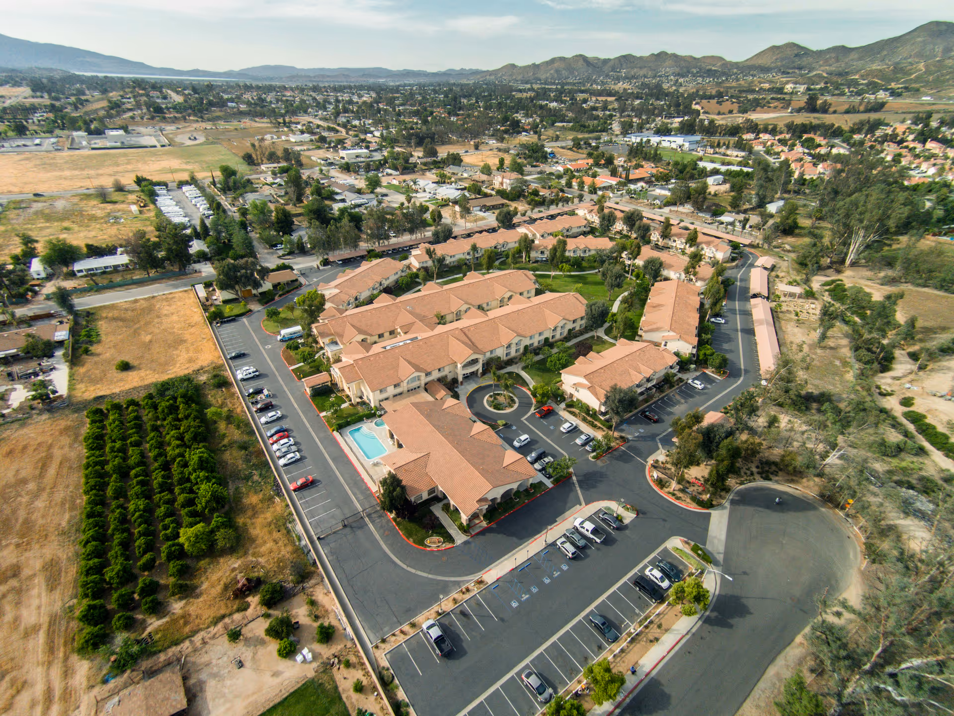 Aerial view of Wildomar Senior Assisted Living facility showing multiple connected buildings with red-tiled roofs, surrounding parking lots with cars, a small swimming pool, landscaped green areas, and nearby fields and residential neighborhoods with mountains in the background.