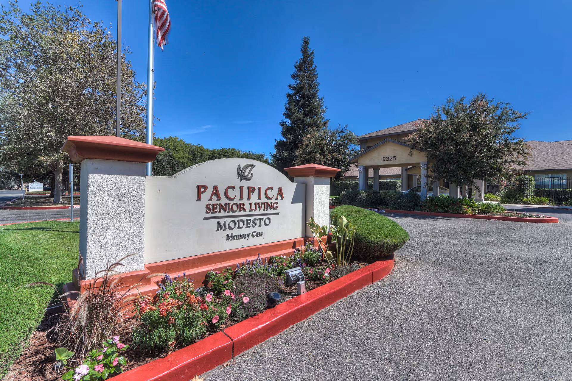 Entrance sign for Pacifica Senior Living Modesto Memory Care with landscaped flowers and bushes, an American flag on a flagpole, and the facility building in the background under a clear blue sky.