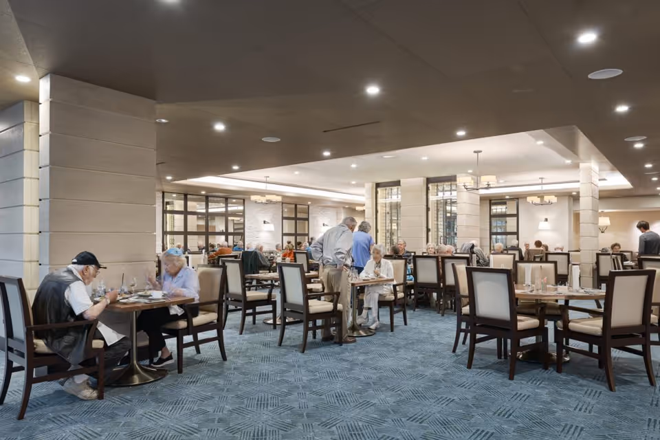 Spacious, well-lit communal dining room with seniors seated at tables and staff assisting at a modern facility.