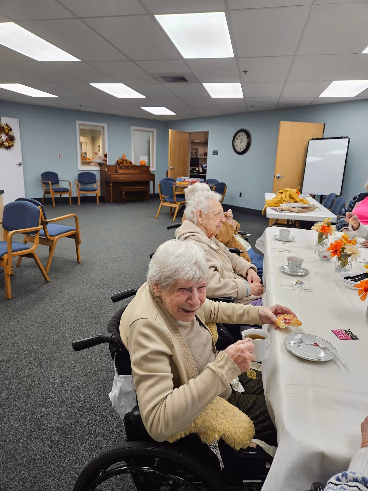 Several elderly women sitting in wheelchairs around a long table in a well-lit room. One woman in the foreground is smiling and holding a cup of tea and a pastry. The table is covered with a white tablecloth and decorated with small flower arrangements. The room has blue walls, several chairs, a piano, a clock on the wall, and a whiteboard in the background.