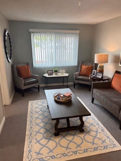 A cozy living room with a gray sofa and two gray armchairs, each with an orange cushion. A wooden coffee table sits on a patterned rug in the center of the room. A small table with framed photos and a lamp is next to the sofa, and a window with vertical blinds lets in natural light. A decorative mirror hangs on the wall to the left.