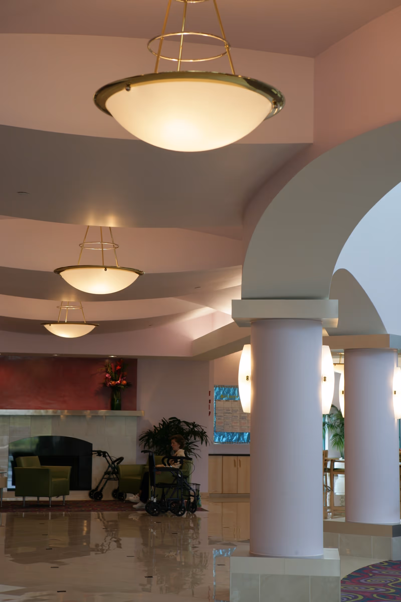 Interior view of a senior living facility lounge area with large white columns, modern ceiling lights, a fireplace with a red accent wall, green chairs, a person sitting in a wheelchair, and plants in the background.