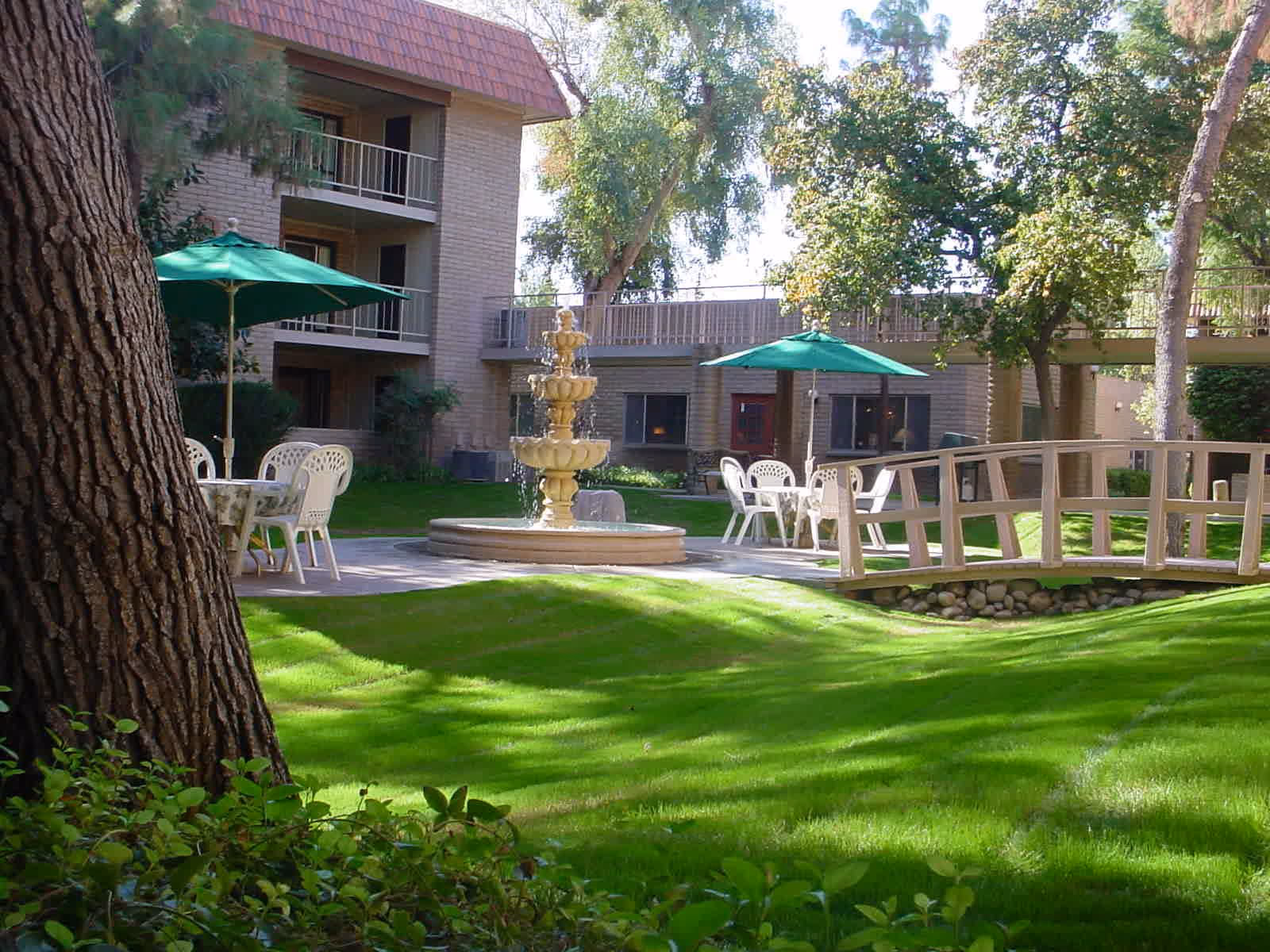 Outdoor courtyard area at Fellowship Square Historic Mesa featuring a green lawn, a multi-tiered water fountain, white plastic tables and chairs with green umbrellas, a small wooden footbridge over a rock-lined stream, and a multi-story building with balconies in the background surrounded by trees and shrubs.