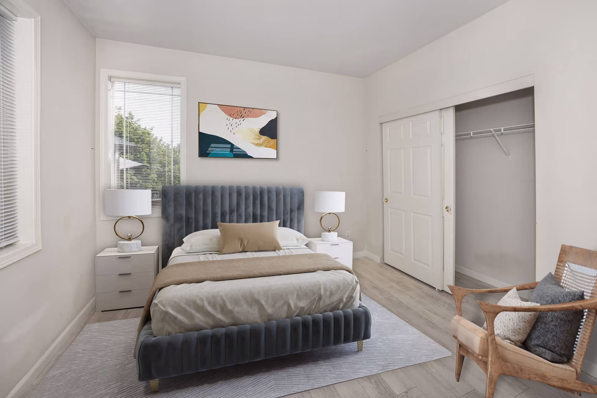 A neatly arranged bedroom with a gray upholstered bed featuring a beige blanket and pillows. There are two white nightstands on either side of the bed, each with a modern lamp. A colorful abstract painting hangs above the bed. The room has light-colored walls, a window with blinds, a closet with sliding doors, and a wooden chair with cushions in the corner.