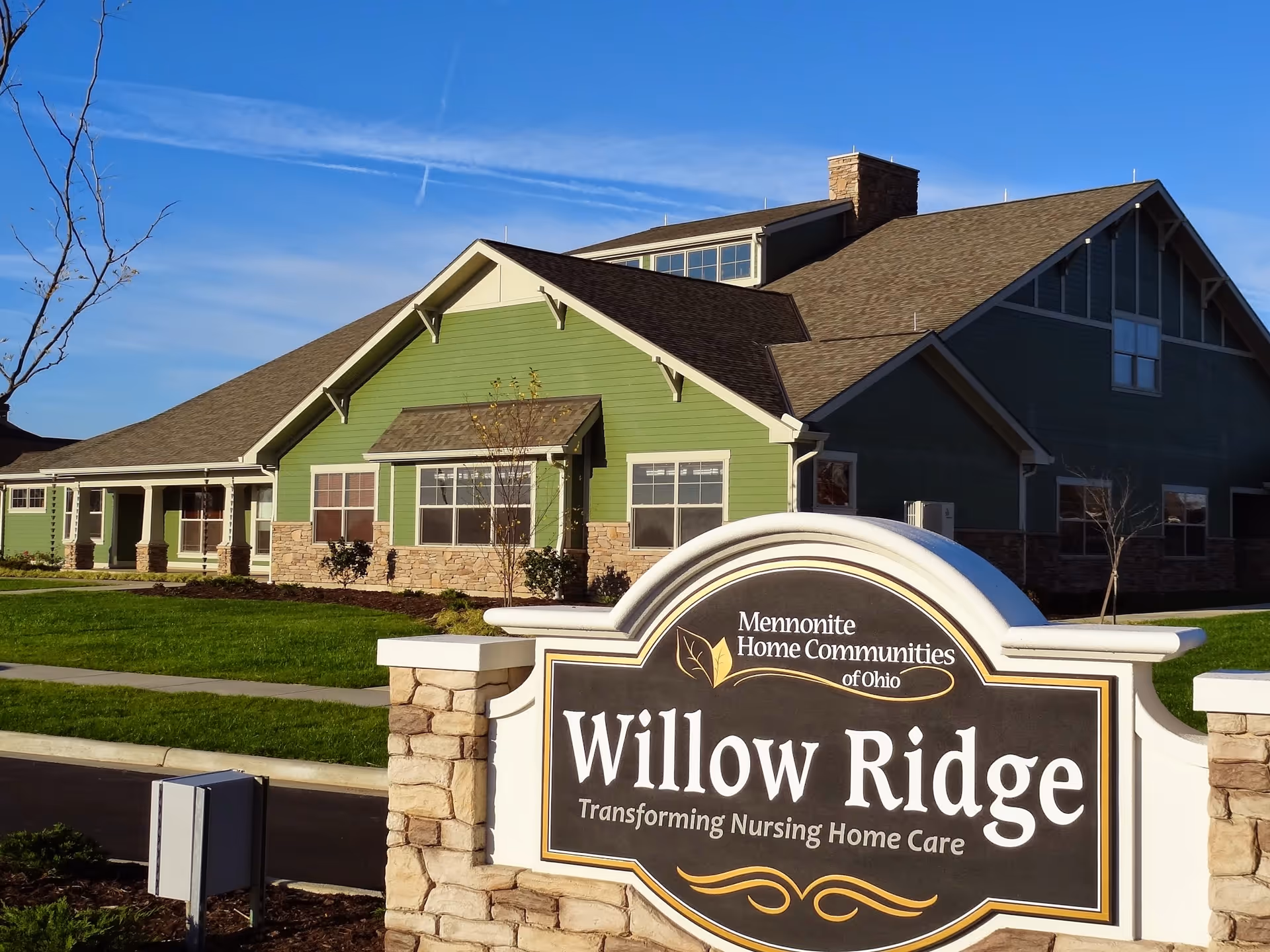 Exterior view of Willow Ridge, a nursing home facility with green siding and stone accents under a clear blue sky. A large sign in front reads 'Willow Ridge, Transforming Nursing Home Care' with the Mennonite Home Communities of Ohio logo above it.