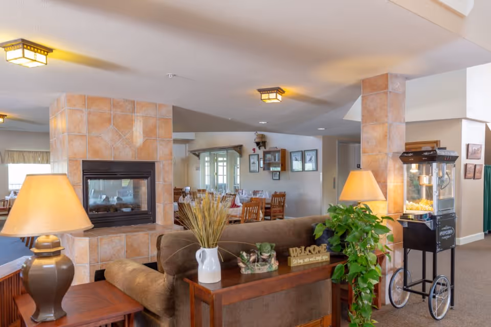 Interior view of a senior living facility common area with a brown couch, two table lamps, a decorative vase with dried plants, a green leafy plant, a popcorn machine on wheels, and a tiled fireplace. In the background, there is a dining area with wooden tables and chairs, and framed pictures on the wall.