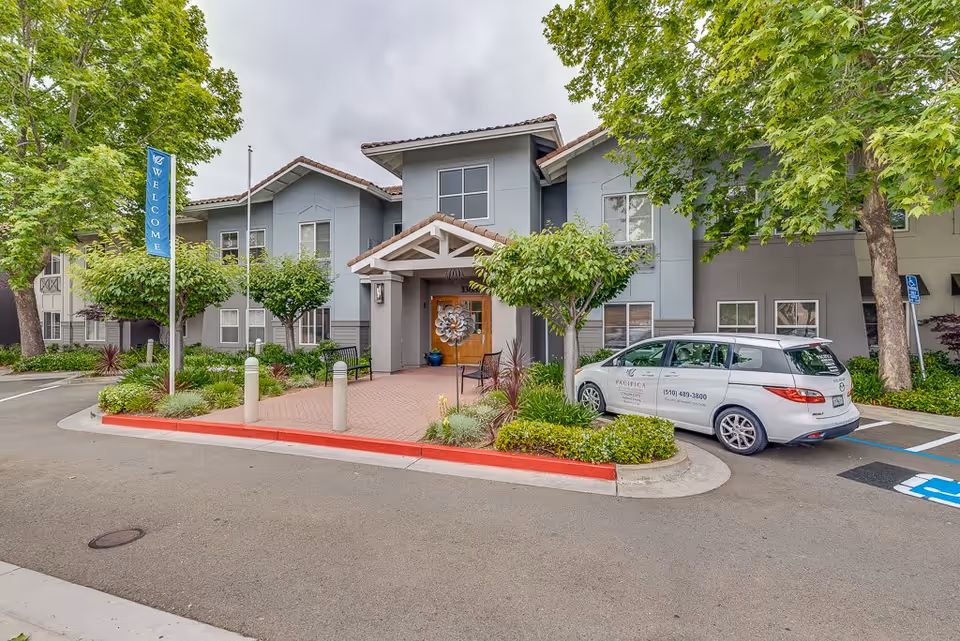 Exterior view of a senior living facility building with a welcoming entrance, surrounded by trees and landscaping. A white vehicle with the Pacifica Senior Living logo and contact information is parked in a handicapped parking spot near the entrance. A blue vertical banner with the word 'WELCOME' is displayed on a pole near the driveway.