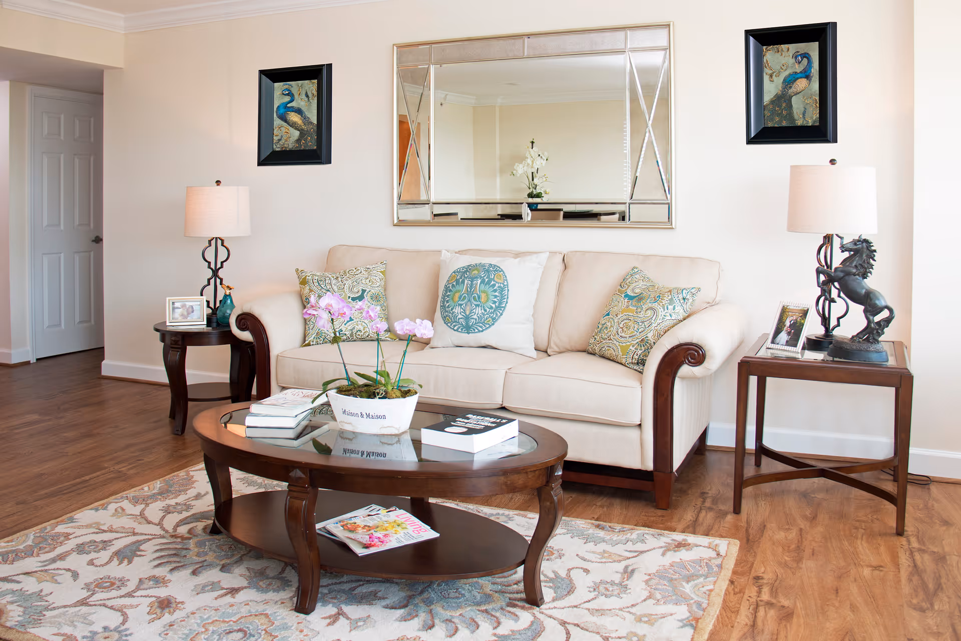 A cozy living room with a beige sofa adorned with decorative pillows, two wooden side tables each holding a lamp and framed photos, a wooden coffee table with books and a potted orchid, a large decorative mirror on the wall, and two framed peacock paintings on either side of the mirror. The room has wooden flooring and a patterned area rug.
