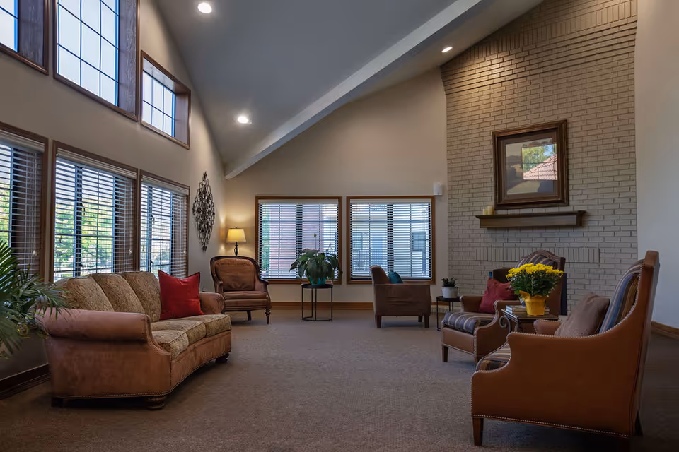 A spacious living room with high vaulted ceilings and large windows allowing natural light to fill the space. The room features a brown sofa with red pillows, several armchairs, a fireplace with a framed picture above it, and a small table with a yellow flower pot. There are plants and decorative items placed around the room, creating a warm and inviting atmosphere.