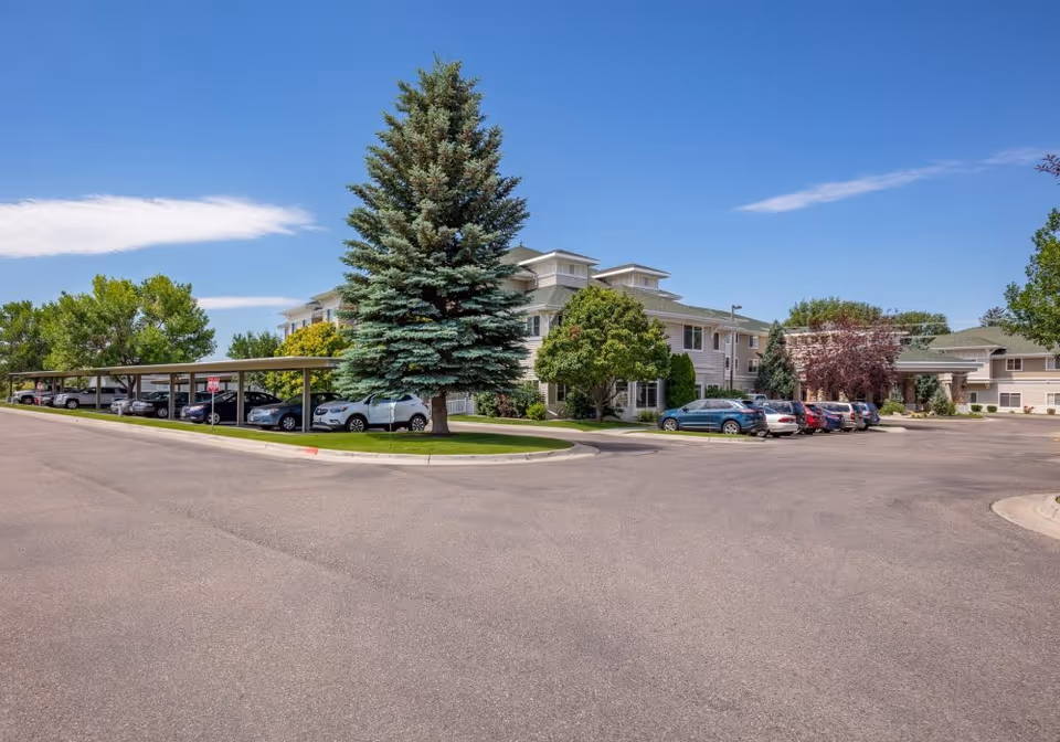Exterior view of a senior living facility with a parking lot, covered parking spaces, several parked cars, and trees under a clear blue sky.