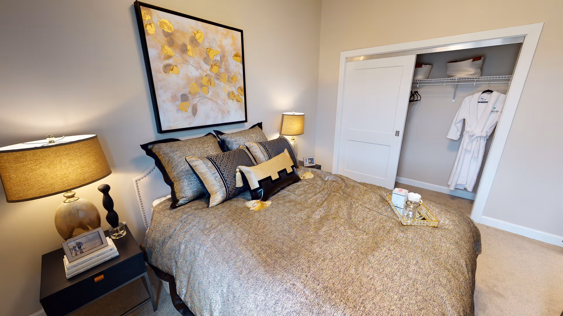 A neatly made bed with multiple decorative pillows in a bedroom. On either side of the bed are nightstands with lamps. Above the bed hangs a framed artwork featuring abstract yellow and gray leaves. To the right, there is an open closet with a white robe hanging inside.