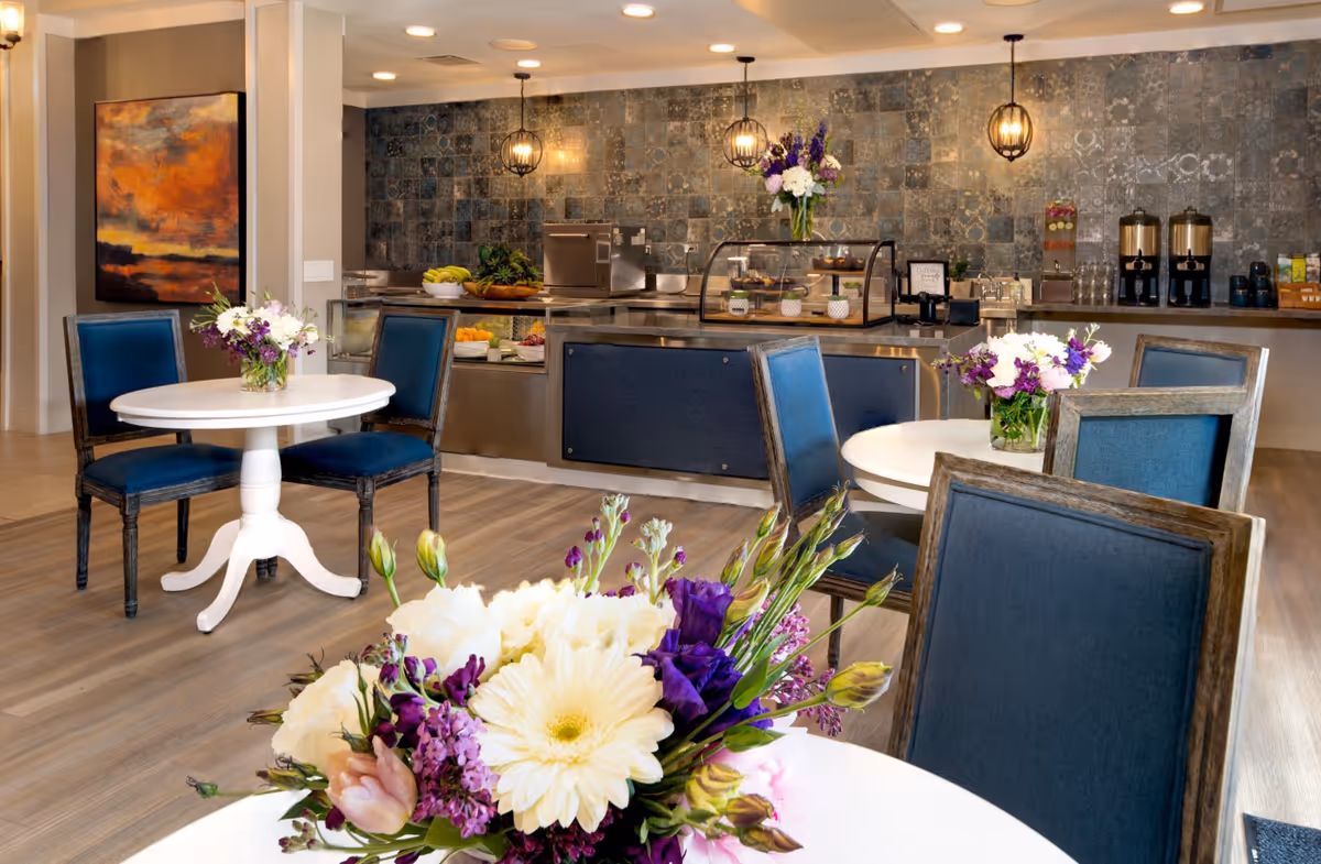 A bright and elegant dining area in La Marea Senior Living featuring round white tables with blue cushioned chairs. Each table has a vase with fresh flowers. In the background, there is a serving counter with food items and beverages, decorative pendant lights, and a textured tile wall.