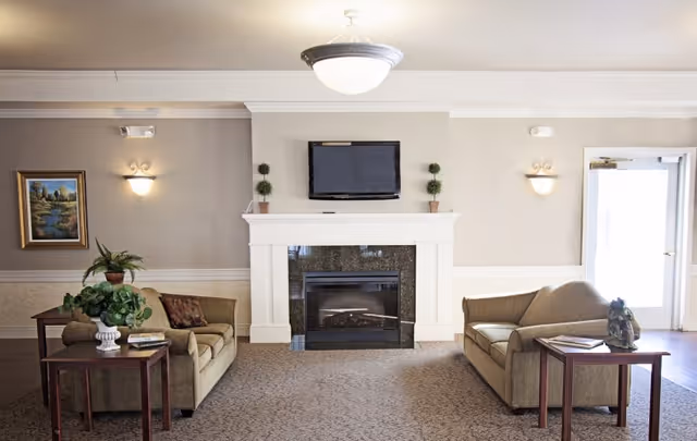 A cozy living room area with two beige sofas facing each other, each accompanied by a wooden side table with decorative items and books. In the center is a fireplace with a black granite surround and a flat-screen TV mounted above it. The room has neutral-colored walls, a ceiling light fixture, wall sconces, a framed landscape painting, and a door letting in natural light.