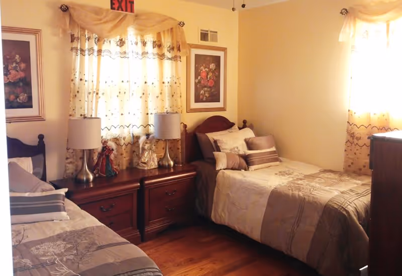 A cozy bedroom with two single beds, each with beige and brown bedding and multiple pillows. Between the beds is a wooden nightstand with two lamps and decorative figurines. The room has light-colored walls, two windows with sheer curtains, and framed floral artwork on the walls.