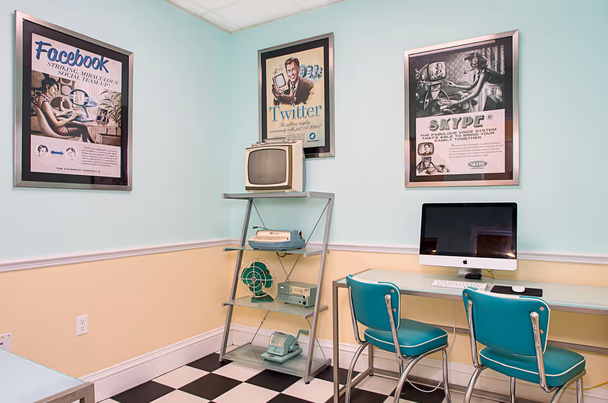 A small room with pastel blue and beige walls featuring three framed vintage-style posters of Facebook, Twitter, and Skype. There is a metal shelving unit holding retro electronics including an old TV, typewriter, fan, and radio. A modern iMac computer sits on a glass desk with two teal retro-style chairs in front of it. The floor has a black and white checkered pattern.