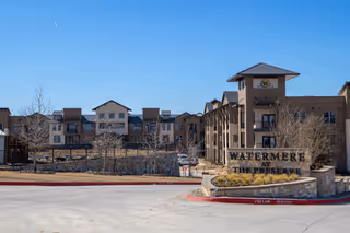 Exterior view of Watermere at the Preserve senior living facility on a clear day, showing a large multi-story building with a stone and brick facade, a prominent sign with the facility's name, and a paved driveway in the foreground.