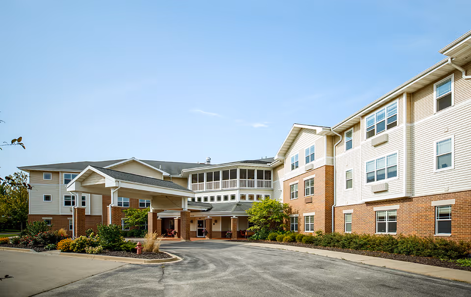 Exterior view of a senior living facility building with a covered entrance, multiple windows, and a landscaped area with shrubs and flowers under a clear blue sky.