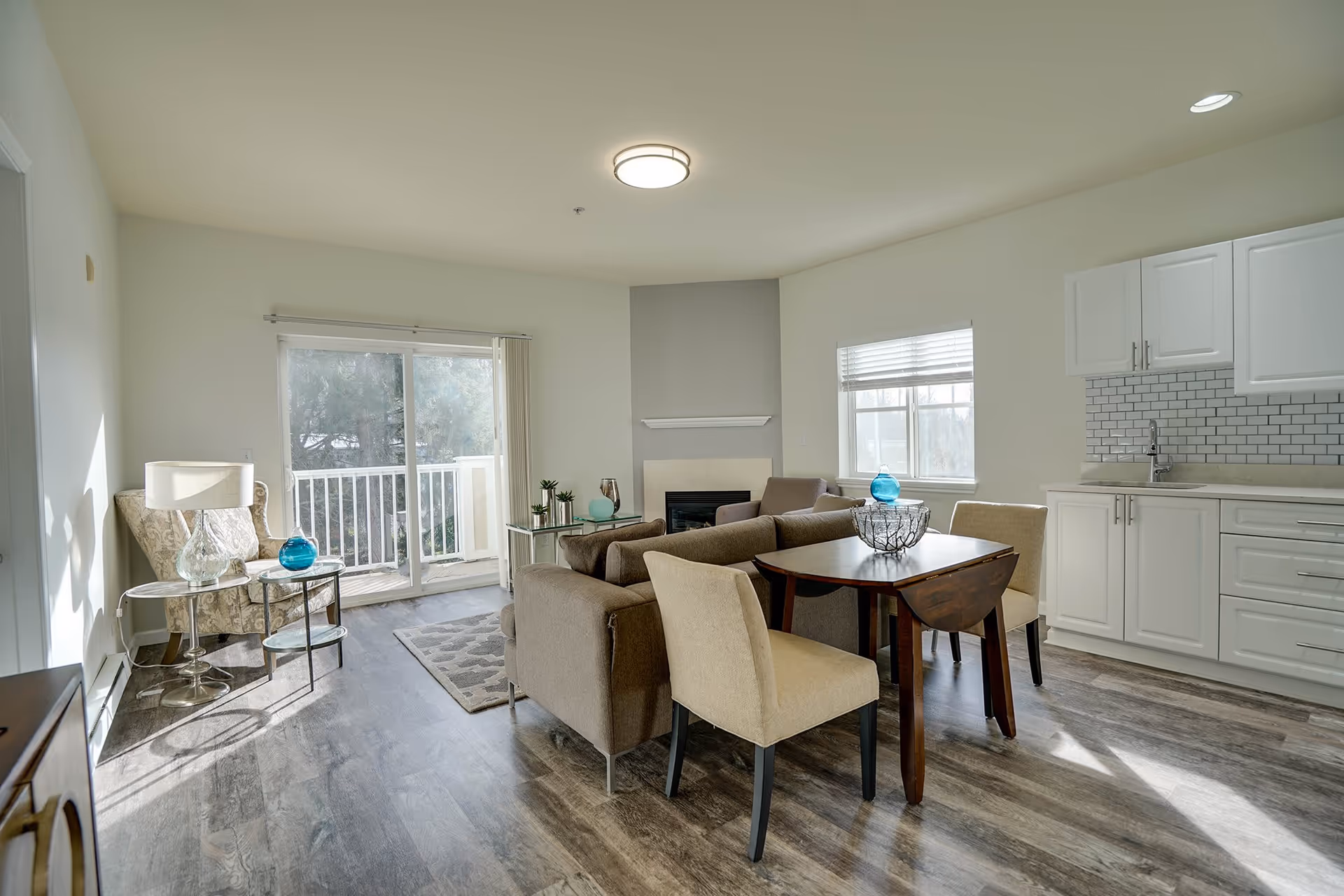 Bright open-plan living room and kitchenette with a sofa, dining table, chairs, sliding glass door to a balcony, and decorative accents.