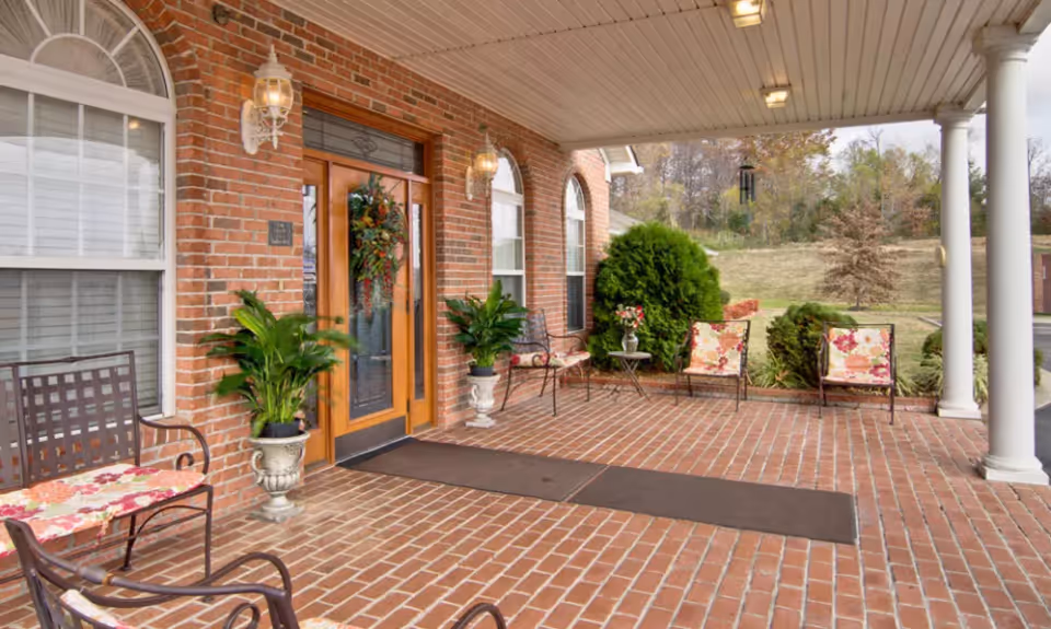 Covered brick patio area outside a building entrance with wooden double doors decorated with a wreath. The patio has several metal chairs with floral cushions, potted plants, and two white columns supporting the roof. In the background, there is a grassy area with trees and shrubs.