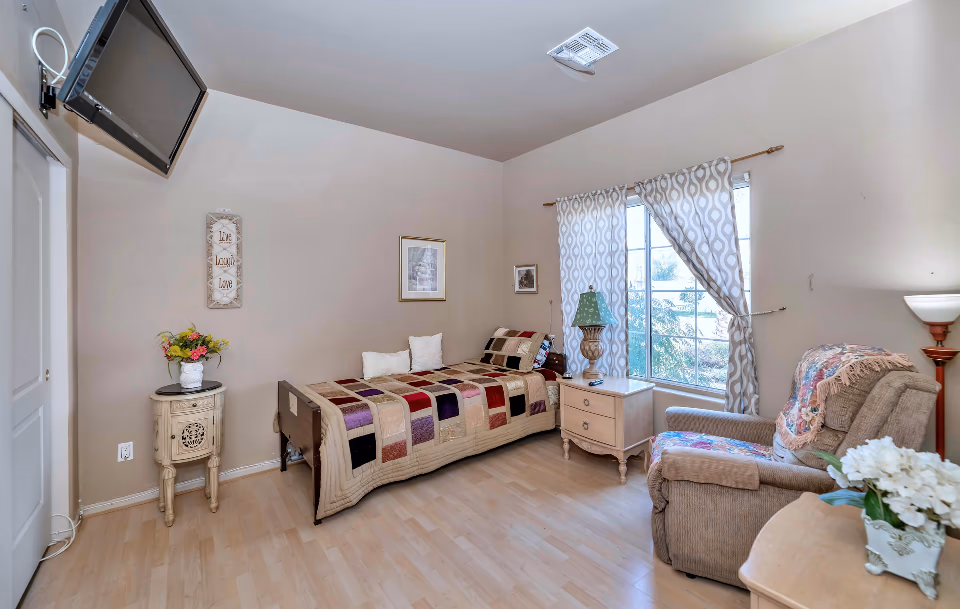 A cozy bedroom in an assisted living facility featuring a single bed with a patchwork quilt, two white pillows, a wooden nightstand with a decorative lamp, a large window with patterned curtains, a comfortable armchair with a colorful throw blanket, a small round table with a flower vase, a wall-mounted TV, and light wood flooring.