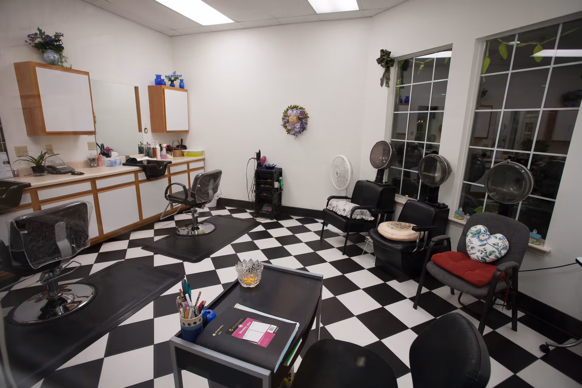 Interior hair salon with black-and-white checkered floor, styling chairs, hooded dryers, mirrors, and a counter with cabinets.