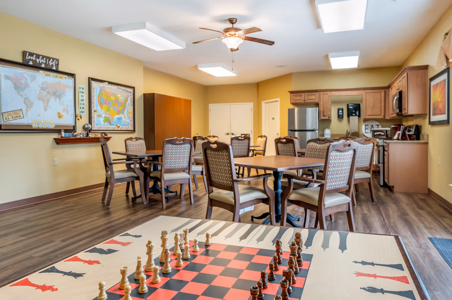 A senior living facility common area with a chess table in the foreground and several wooden tables and chairs arranged in the background. The room has a ceiling fan, bright overhead lights, and a kitchen area with wooden cabinets, a refrigerator, microwave, and coffee maker. There are two framed maps on the wall and a wooden floor.