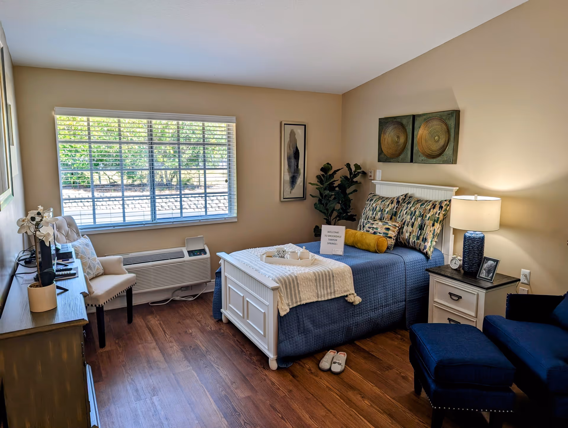 A cozy bedroom in a senior living facility with a white wooden bed frame, blue bedding, and multiple patterned pillows. A small tray with a teapot and cups sits on the bed along with a sign that reads 'Welcome to Brookdale Tarpon Springs!'. The room has wooden flooring, a window with blinds letting in natural light, a beige armchair, a blue armchair with matching ottoman, a nightstand with a lamp, clock, and framed photo, and decorative wall art above the bed.