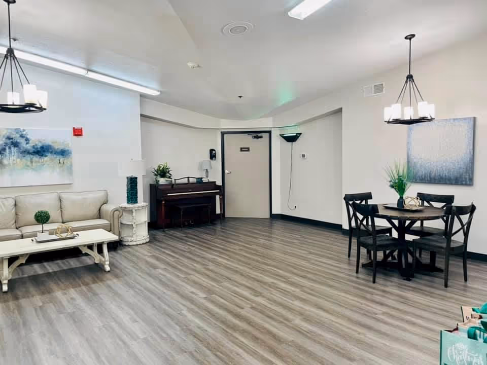 A spacious, well-lit common area with light wood flooring and white walls. The room features a beige sofa with a white coffee table in front, a dark wooden piano against the back wall, and a round dining table with four black chairs. Two modern chandeliers hang from the ceiling, and there are two pieces of artwork on the walls. A small plant and decorative items are placed on the tables and piano.