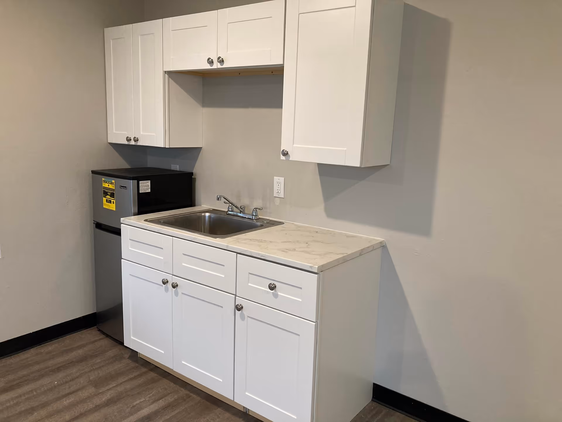 Small kitchenette area with white cabinets above and below a stainless steel sink, a marble-patterned countertop, and a compact stainless steel refrigerator in the corner.