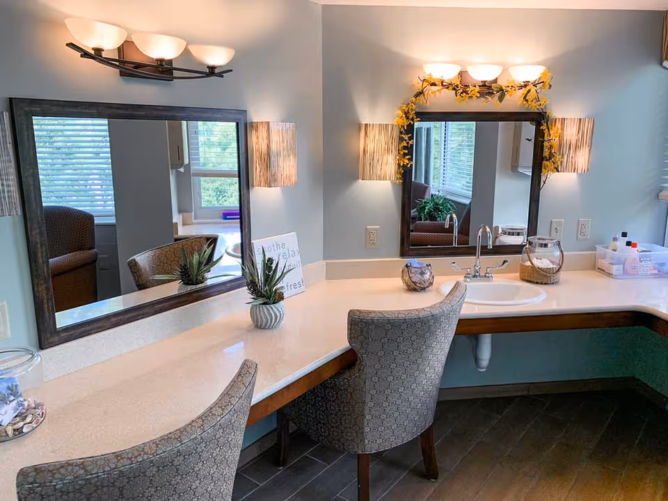 Well-lit vanity area with two mirrors, upholstered chairs, a countertop sink, and decorative plants and lights.