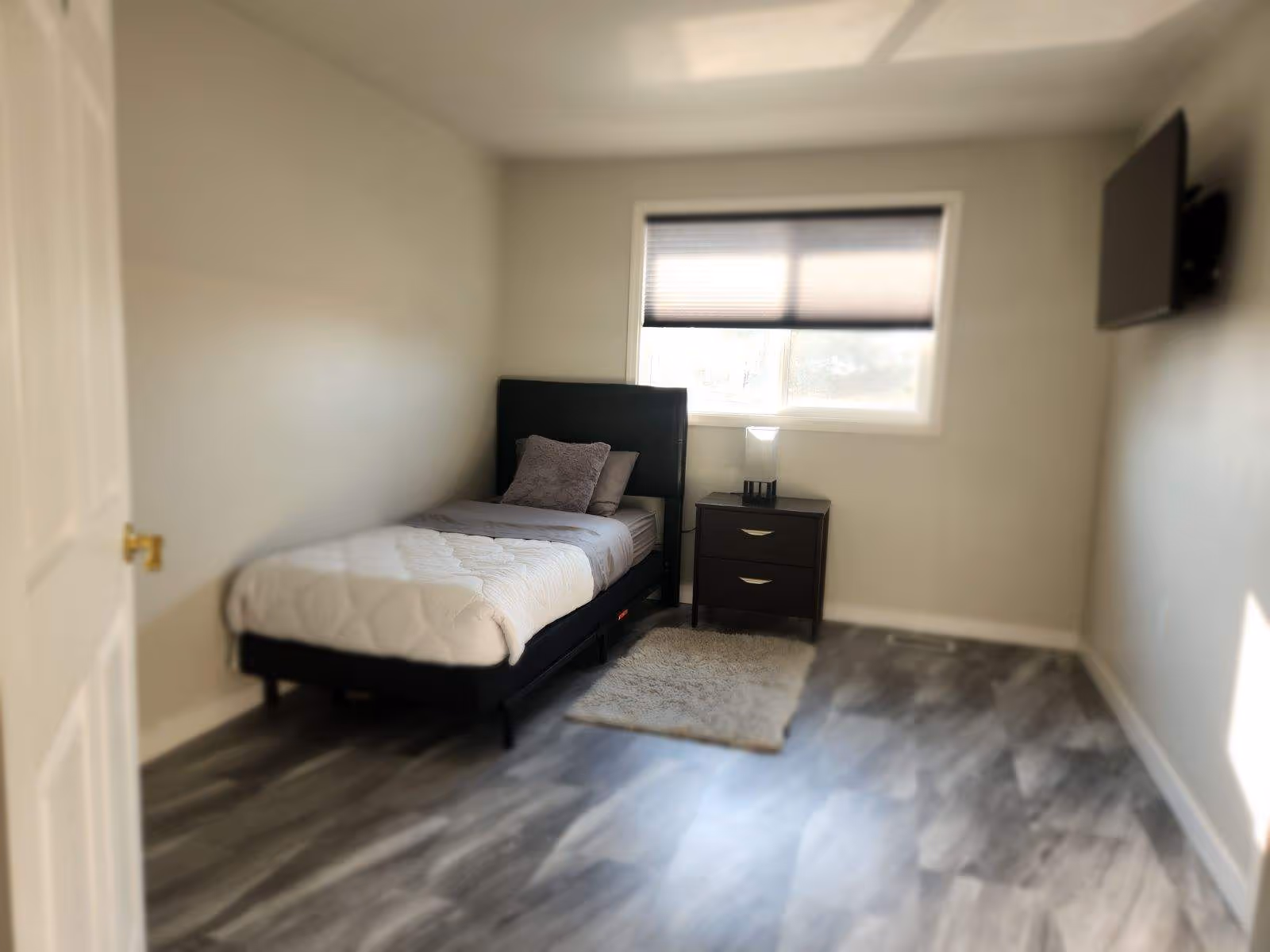 A simple bedroom with a single bed against the left wall, a dark headboard, and two pillows. Next to the bed is a small dark nightstand with two drawers and a lamp on top. A window with a partially closed blind is above the nightstand. A small rug is placed on the floor beside the bed. A flat-screen TV is mounted on the right wall. The room has light-colored walls and wood-patterned flooring.