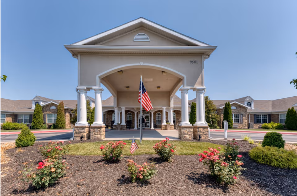 Front exterior view of a senior living facility with a covered entrance supported by white columns, an American flag in the center, and landscaped flower beds with red flowers in front. The building has a beige facade with stone accents and multiple windows under a clear blue sky.
