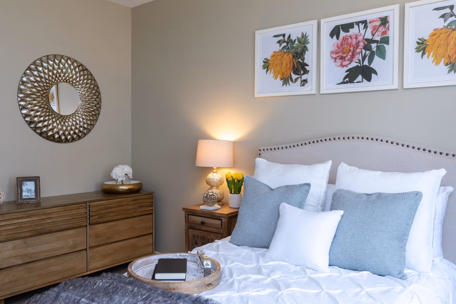 A cozy bedroom with a neatly made bed featuring white and light blue pillows. A wooden nightstand with a decorative lamp and a vase of yellow tulips sits beside the bed. Above the bed are three framed floral prints. A wooden dresser with a round decorative mirror and a small framed photo is against the adjacent wall.