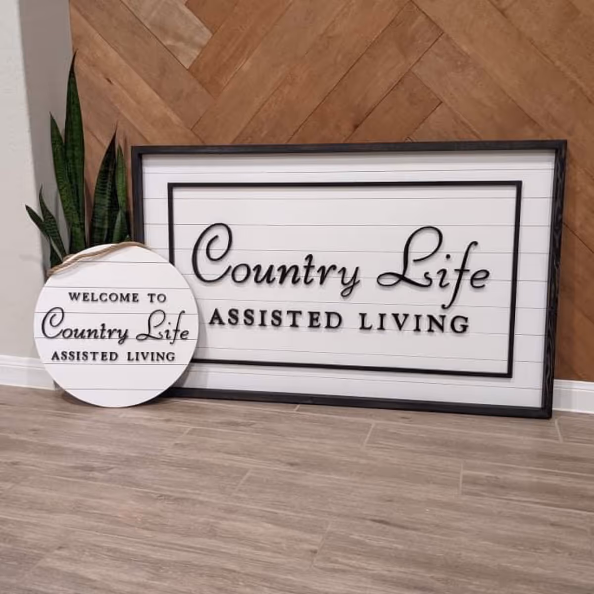 Two decorative signs leaning against a wooden herringbone patterned wall on a wooden floor. One large rectangular sign reads 'Country Life Assisted Living' and a smaller round sign reads 'Welcome to Country Life Assisted Living'. There is a green plant behind the signs.