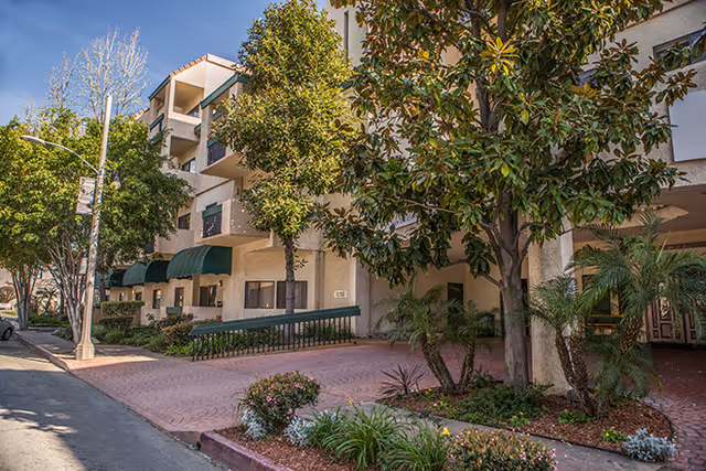 Exterior view of a multi-story residential building with balconies and green awnings over the windows. The building is surrounded by trees and landscaped plants along a brick-paved walkway and driveway under a clear blue sky.