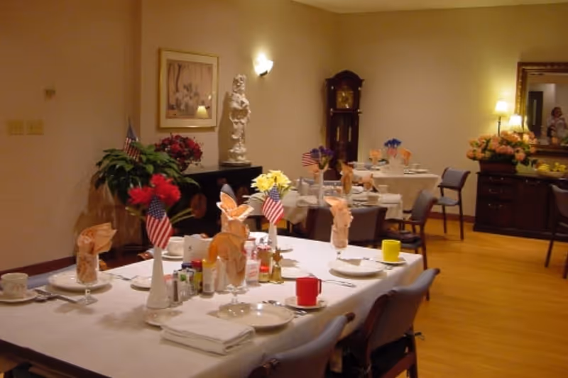 A dining room with tables set for a meal, decorated with small American flags, flowers in vases, folded napkins, and various condiments. The room has wooden flooring, a grandfather clock, a sideboard with a large mirror, and soft lighting from wall sconces and lamps.