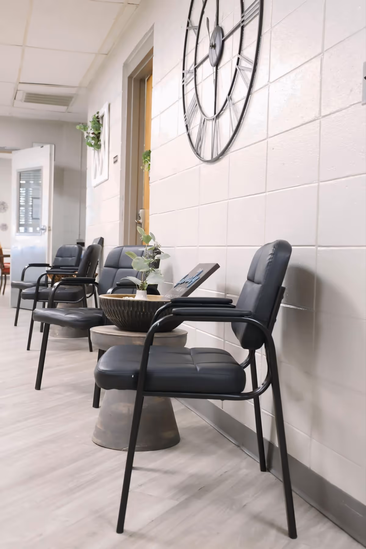 Row of black chairs and small tables with potted plants along an interior hallway decorated with a large wall clock.