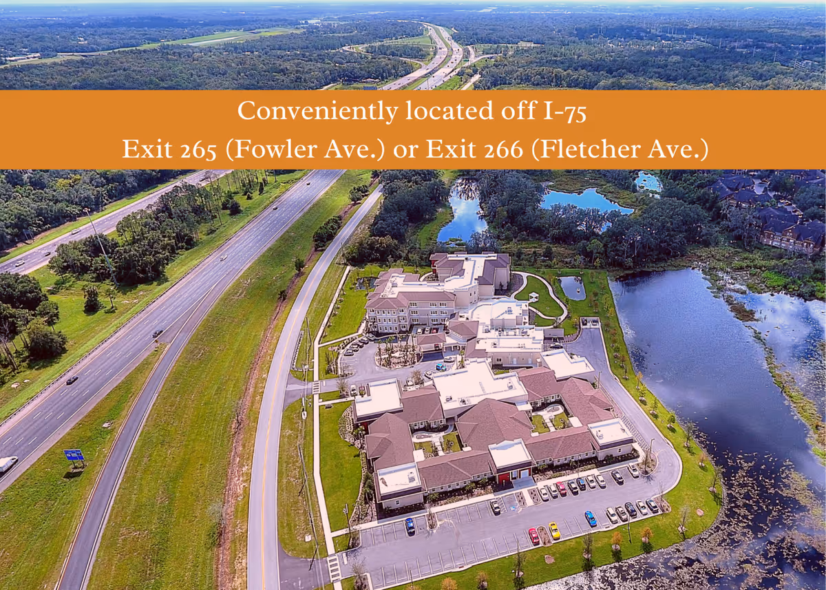 Aerial view of the Promise Pointe Senior Living campus beside a highway and a lake with an orange banner giving nearby I-75 exit information.