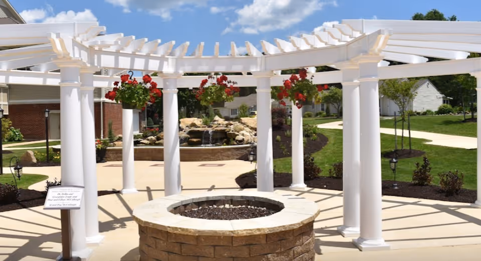 Outdoor patio area with a white pergola structure supported by columns, hanging flower baskets with red flowers, a circular stone fire pit in the center, and a landscaped garden with a small waterfall feature in the background under a blue sky with some clouds.