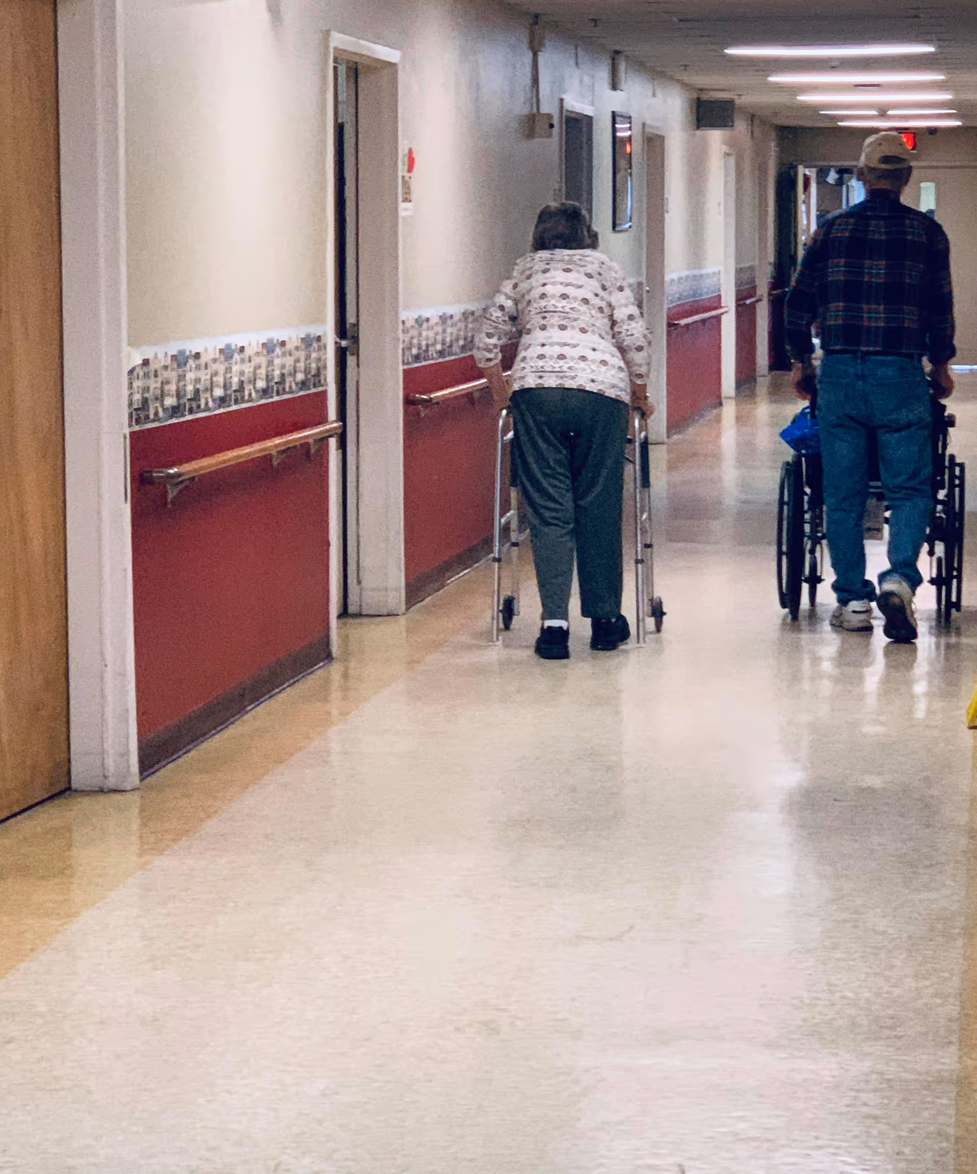 Two elderly individuals walking down a long hallway in a senior living facility. One person is using a walker, and the other is pushing a wheelchair. The hallway has red and beige walls with handrails and several doors along the sides.