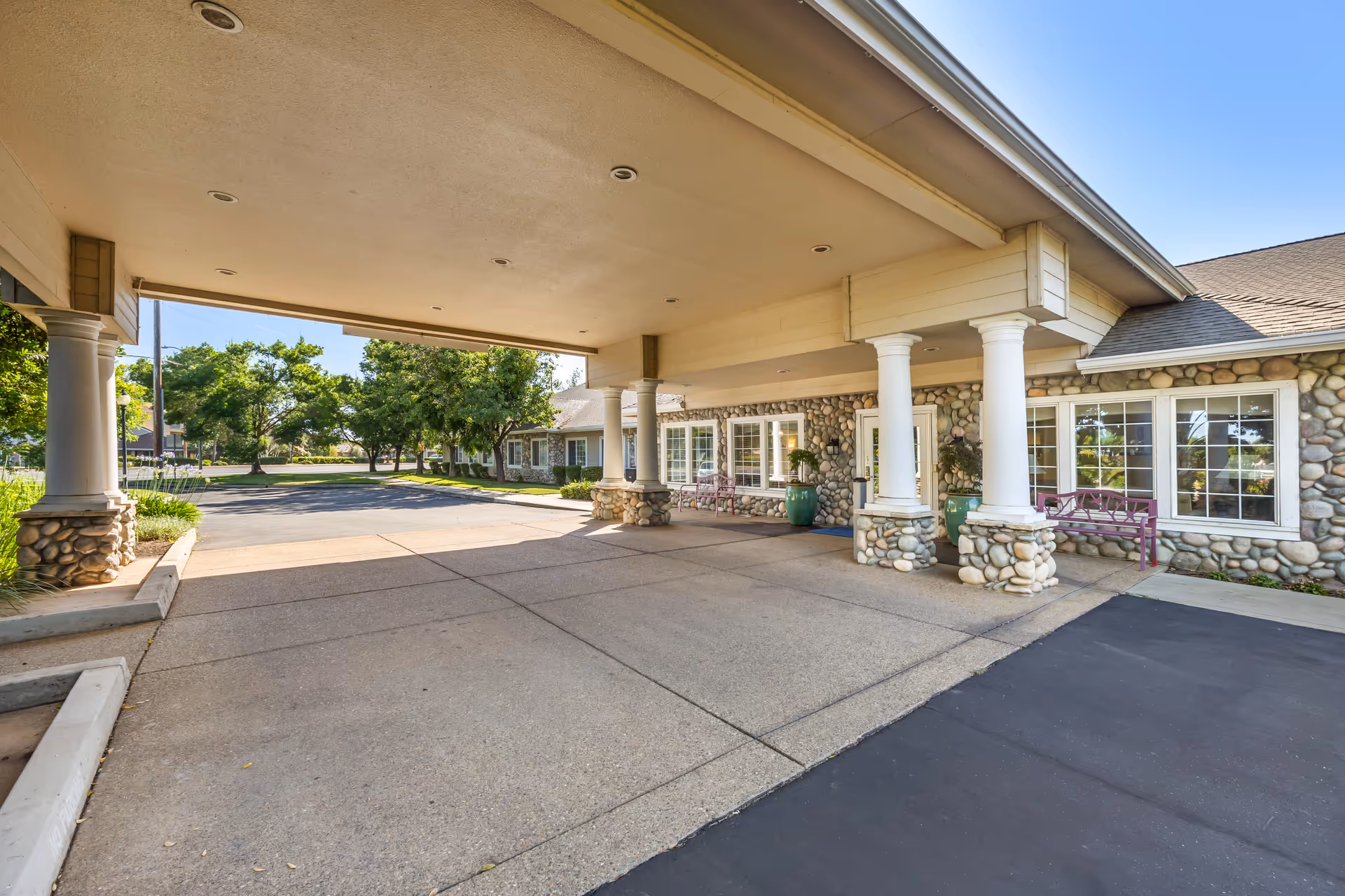 Covered driveway entrance to a building with stone pillars and walls, large windows, and benches outside. Trees and a clear blue sky are visible in the background.