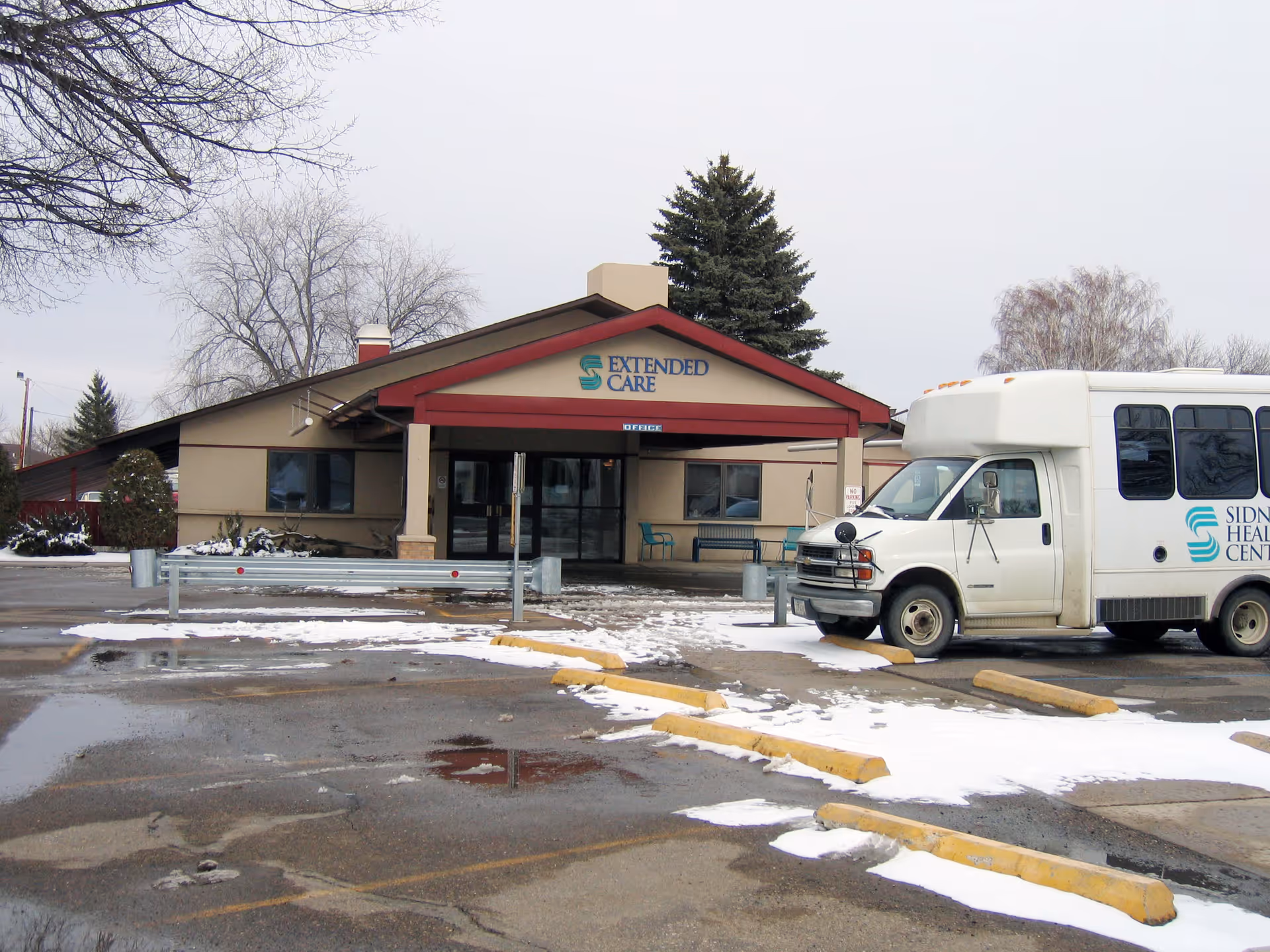 Front entrance of Sidney Health Center Extended Care with a parked shuttle bus and a snowy parking lot.