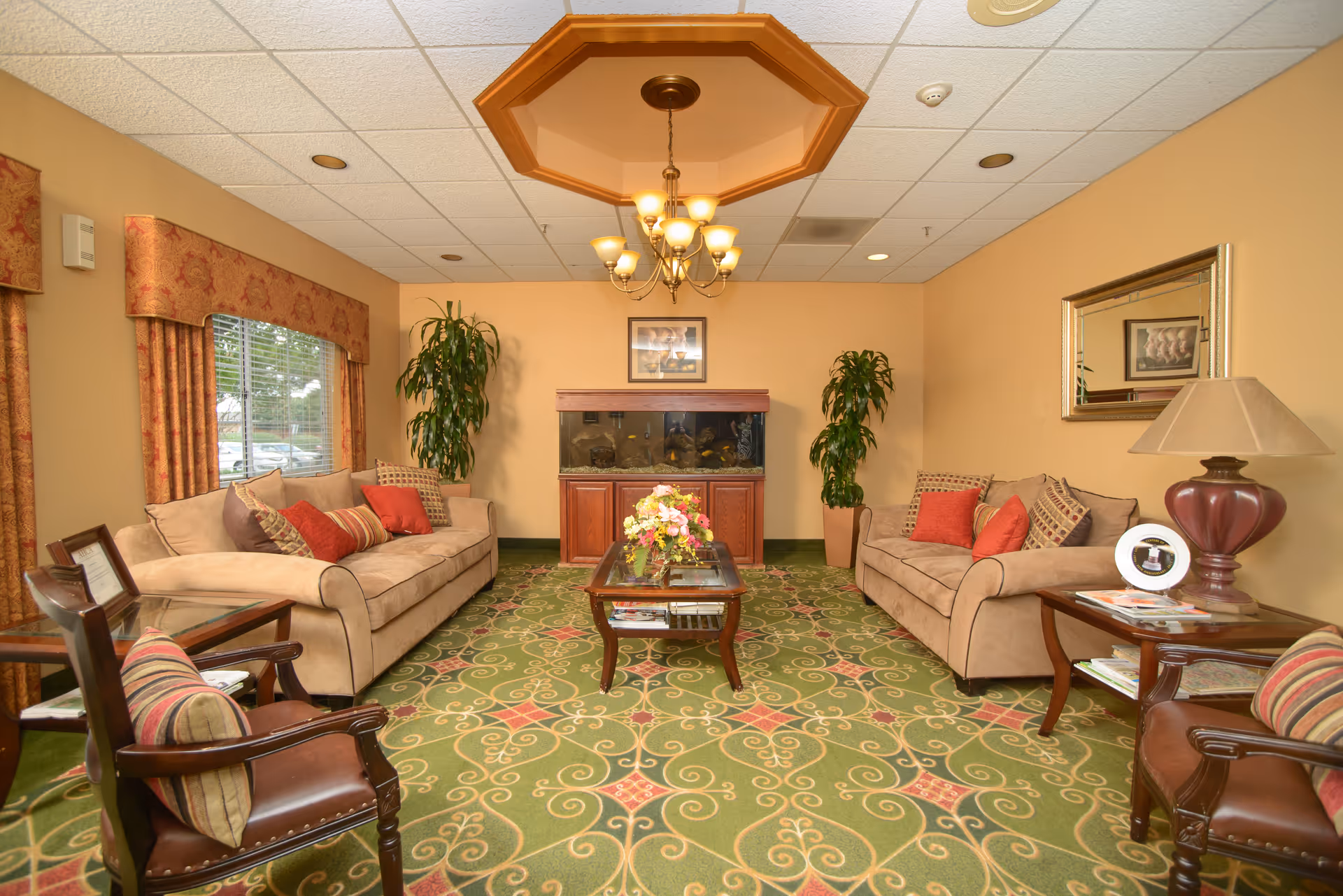 Comfortable furnished sitting room with two sofas, armchairs, a coffee table, an aquarium against the back wall, plants and a chandelier.