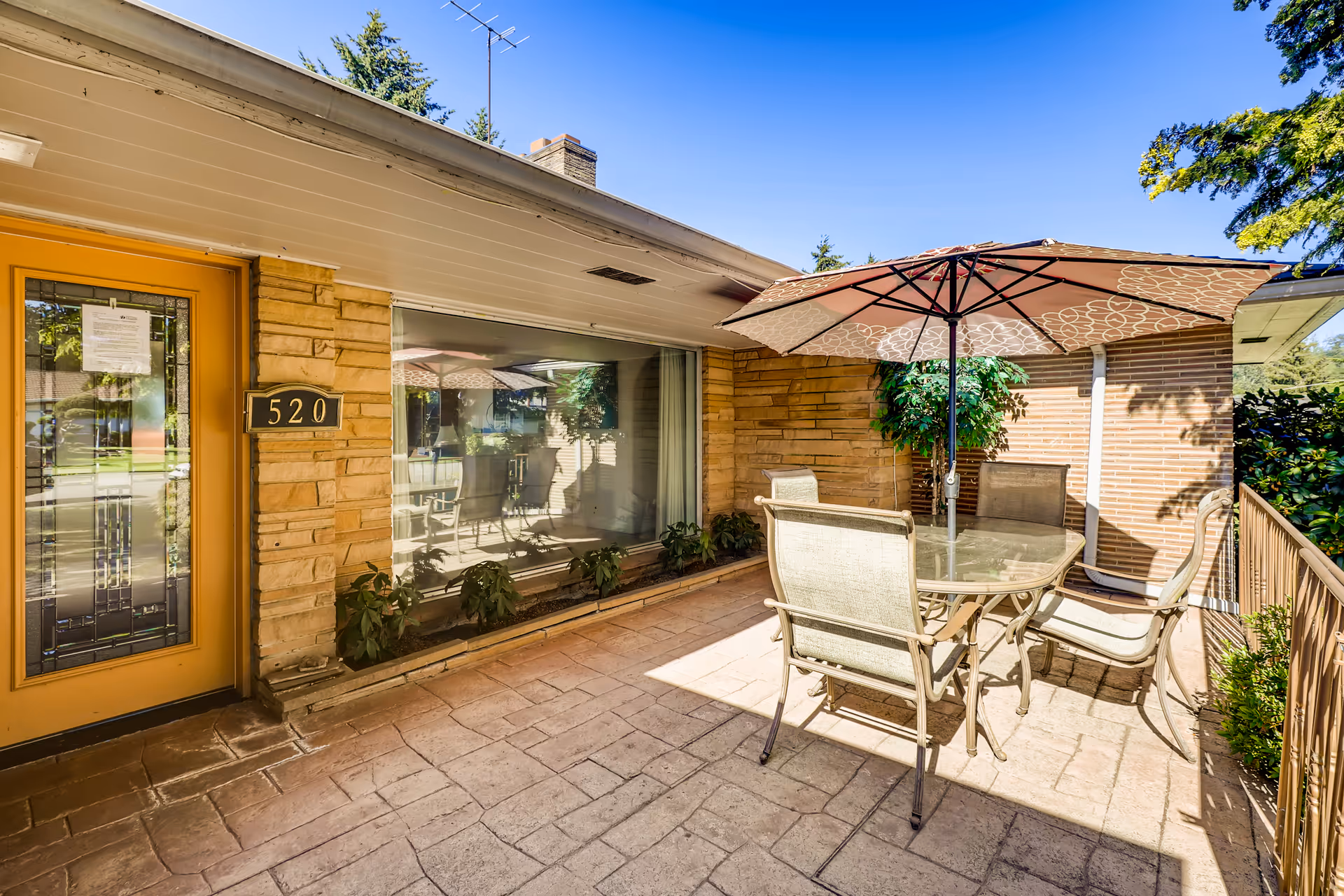 Outdoor patio area at New Options Renton AFH featuring a glass-top table with four chairs and a large patterned umbrella. The patio is paved with stone tiles and is adjacent to a building with a yellow door numbered 520 and a large window reflecting the patio furniture. There are plants along the base of the building and trees visible in the background under a clear blue sky.